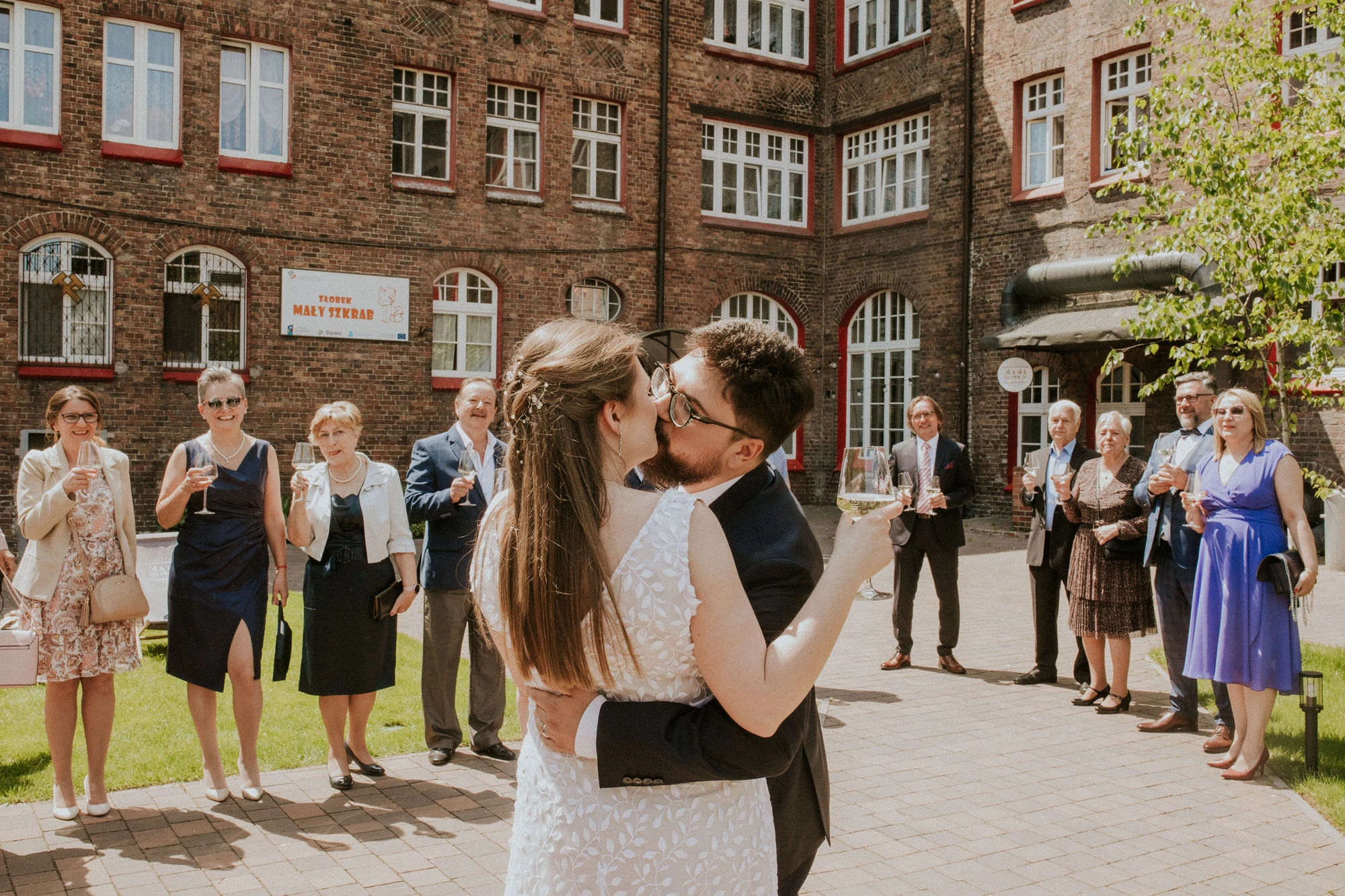 Para młoda otoczona weselnymi gośćmi przed budynkiem.