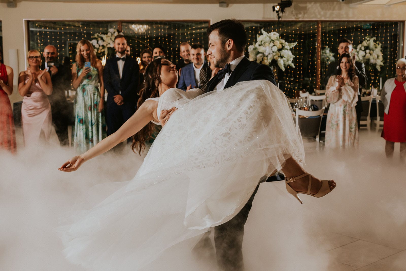 Para młoda tańczy z dymem i gośćmi w tle.