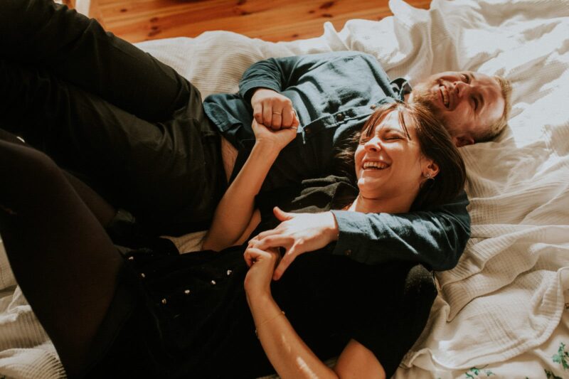 Para śmiejąca się na łóżku, trzymająca się za ręce.