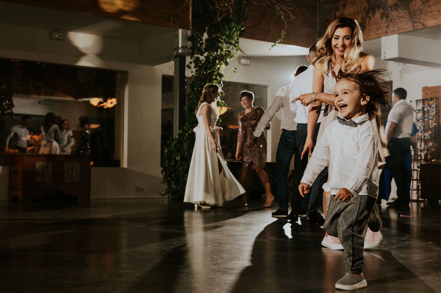 Dziecko tańczy na weselu, uśmiechnięci goście w tle.