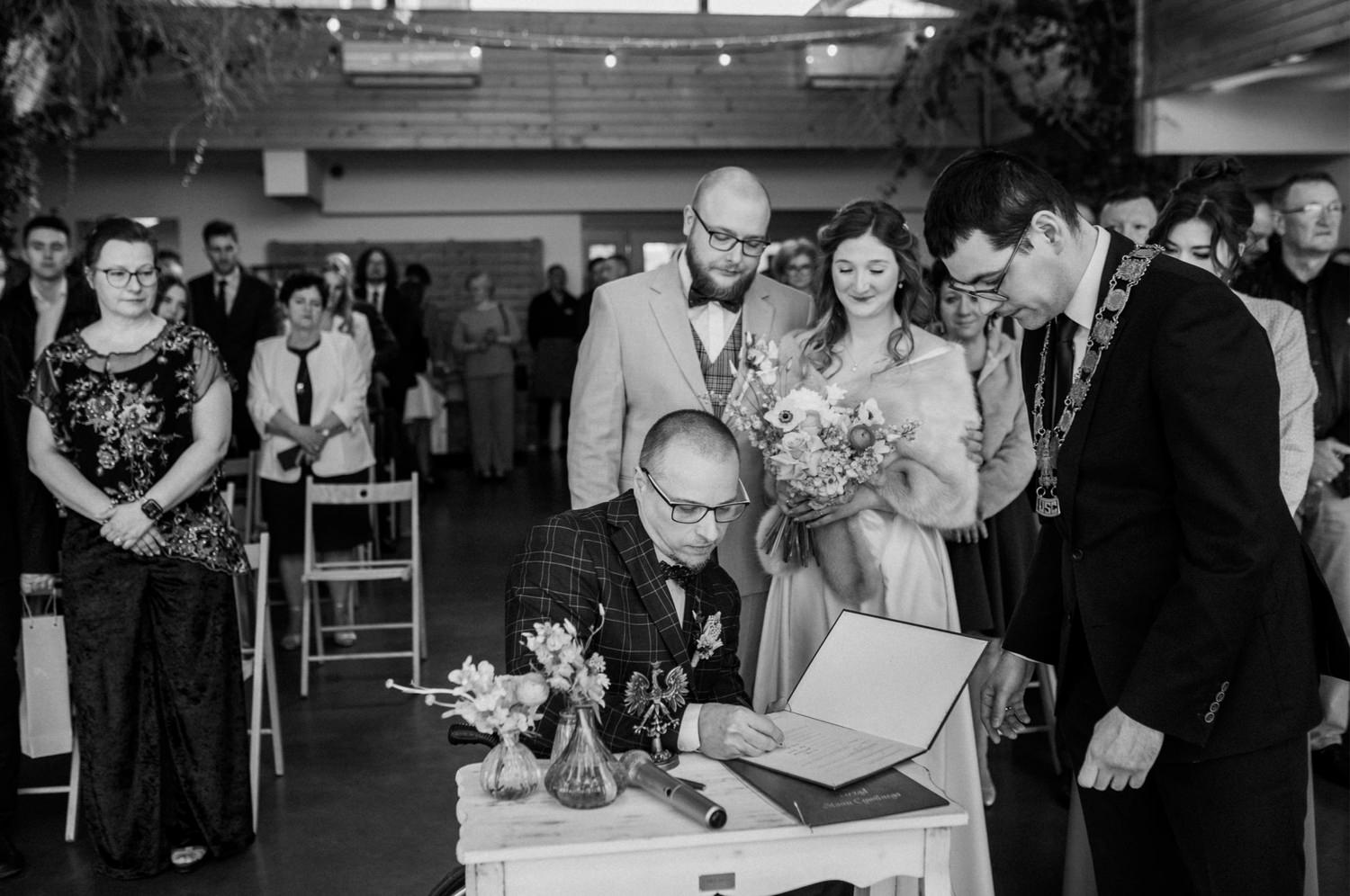 Ślub cywilny, podpisywanie dokumentów, goście w tle.