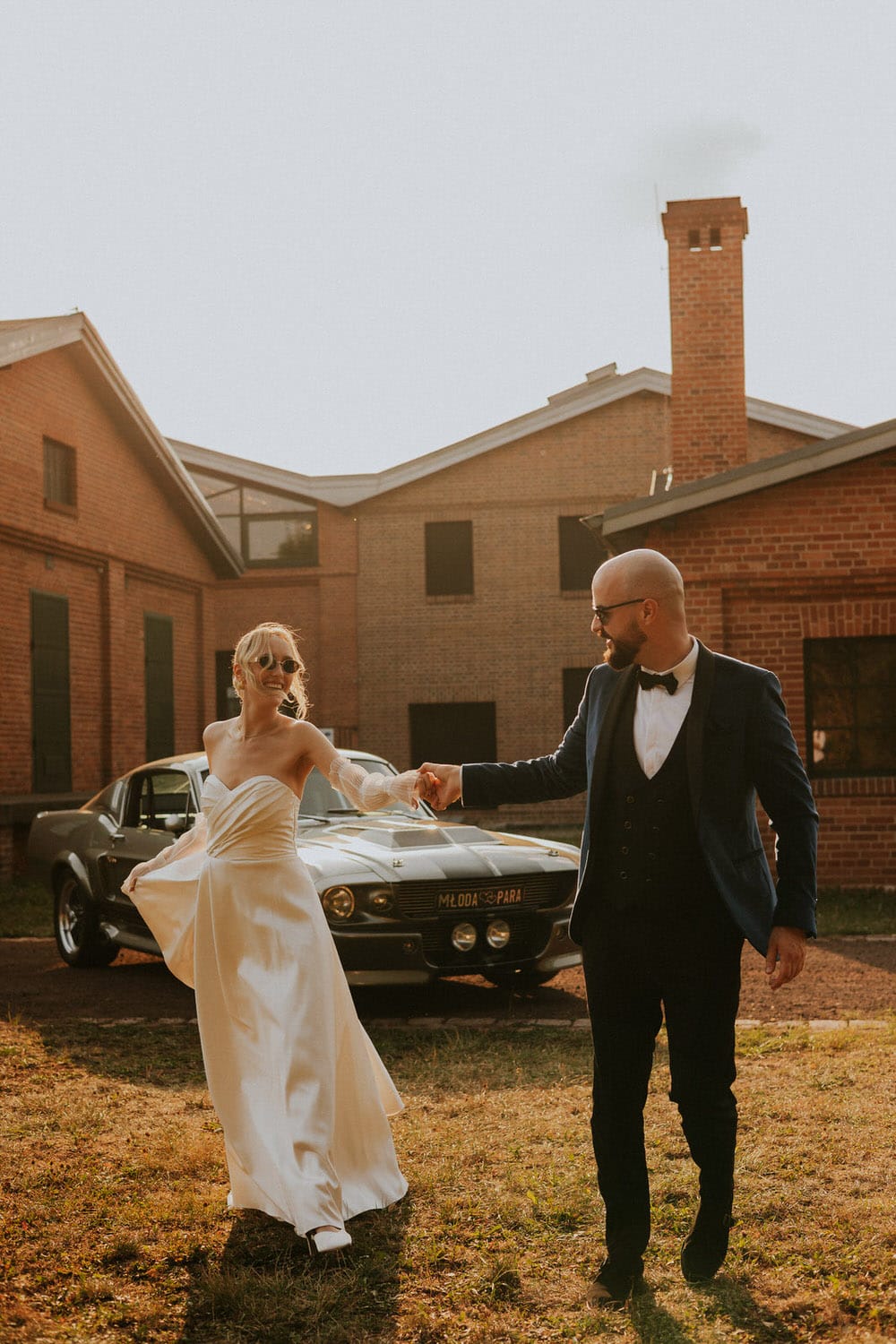 Para młoda tańczy przed zabytkowym samochodem, w tle ceglana chata.